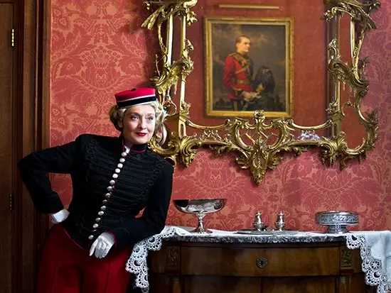 Fiona Harrison in a red military-style jacket and hat in an ornate room